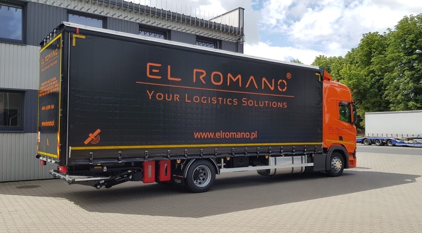 Pomarańczowa ciężarówka z czarną naczepą firmy El Romano z Dominowa, zaparkowana na brukowanym placu, widok z tyłu z boku, w tle budynek i zieleń.