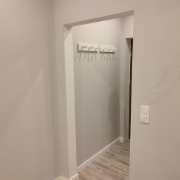 Mp projekt - Szare ściany przedpokoju z drewnopodobną podłogą, białe wieszaki na ścianie i włącznik światła po prawej stronie.