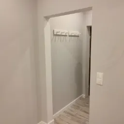 Szare ściany przedpokoju z drewnopodobną podłogą, białe wieszaki na ścianie i włącznik światła po prawej stronie.