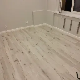 Jasne panele podłogowe ułożone w pustym, świeżo pomalowanym pokoju z szarą ścianą, widoczne gniazdka elektryczne i listwy przypodłogowe.