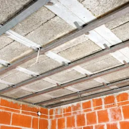 Montaż sufitu podwieszanego z płyt kartonowo-gipsowych na metalowej konstrukcji, widoczne ocieplenie i ceglana ściana w tle.