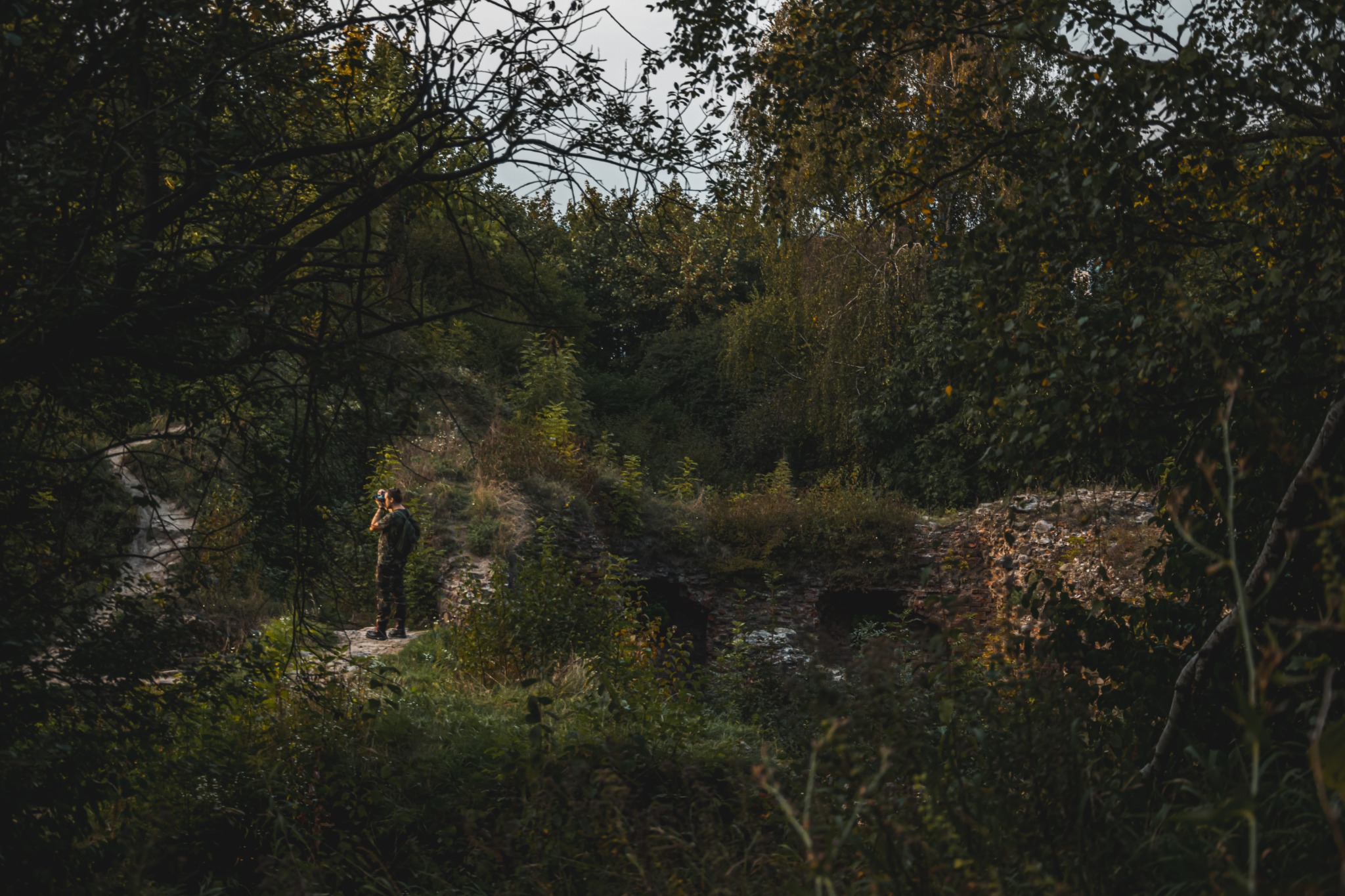 Osoba w kamuflażu z aparatem fotograficznym stojąca na ścieżce w zaroślach, fotografująca krajobraz.