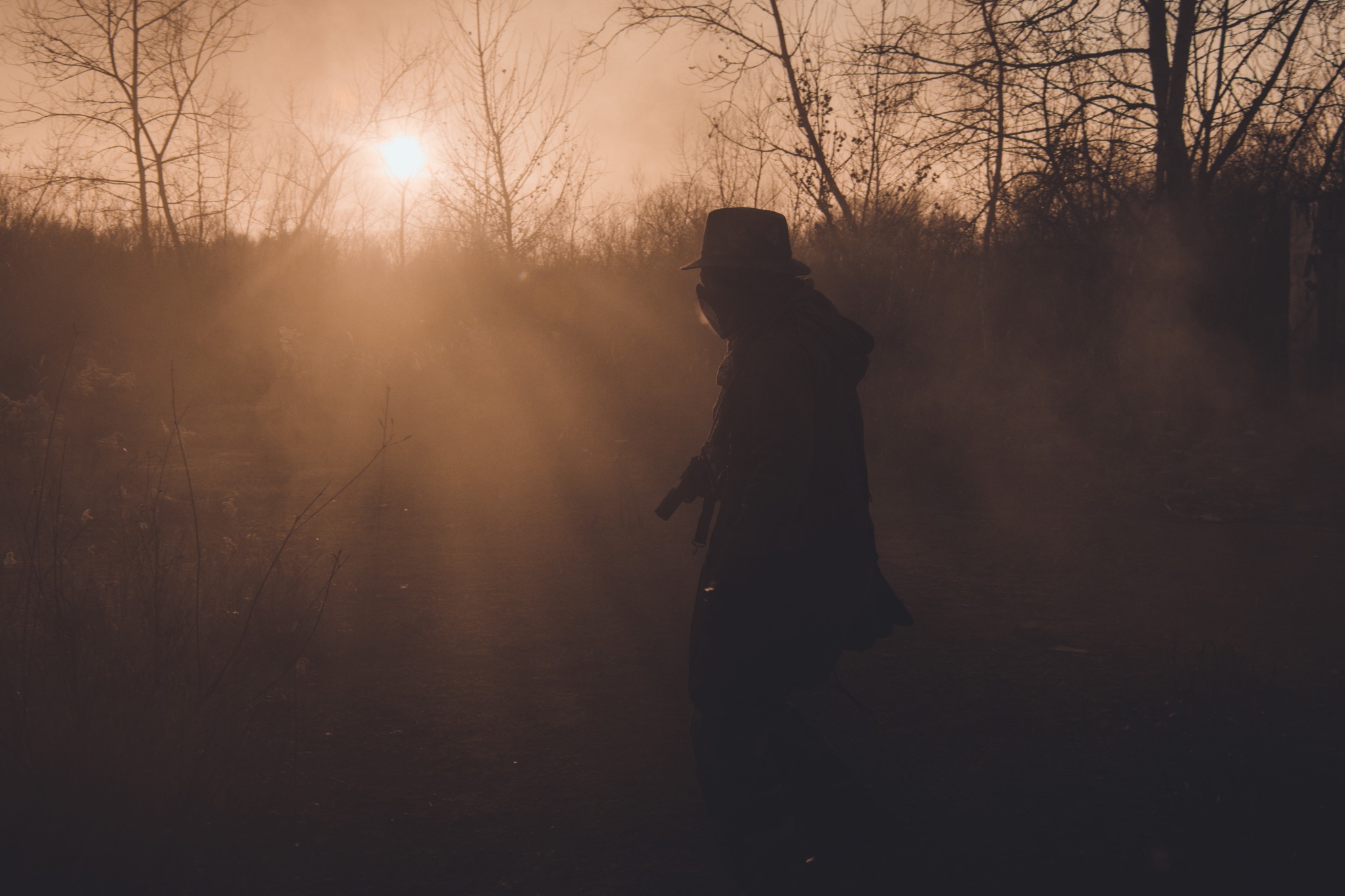 Sylwetka osoby w kapeluszu i płaszczu, trzymającej przedmiot przypominający broń, na tle zalesionego terenu spowitego mgłą o zachodzie słońca.