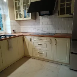 Jasna kuchnia z kremowymi szafkami, drewnianym blatem i białymi kafelkami typu metro, czarny okap nad płytą grzewczą, widoczne szklane witryny szafek g&oacute;rnych.