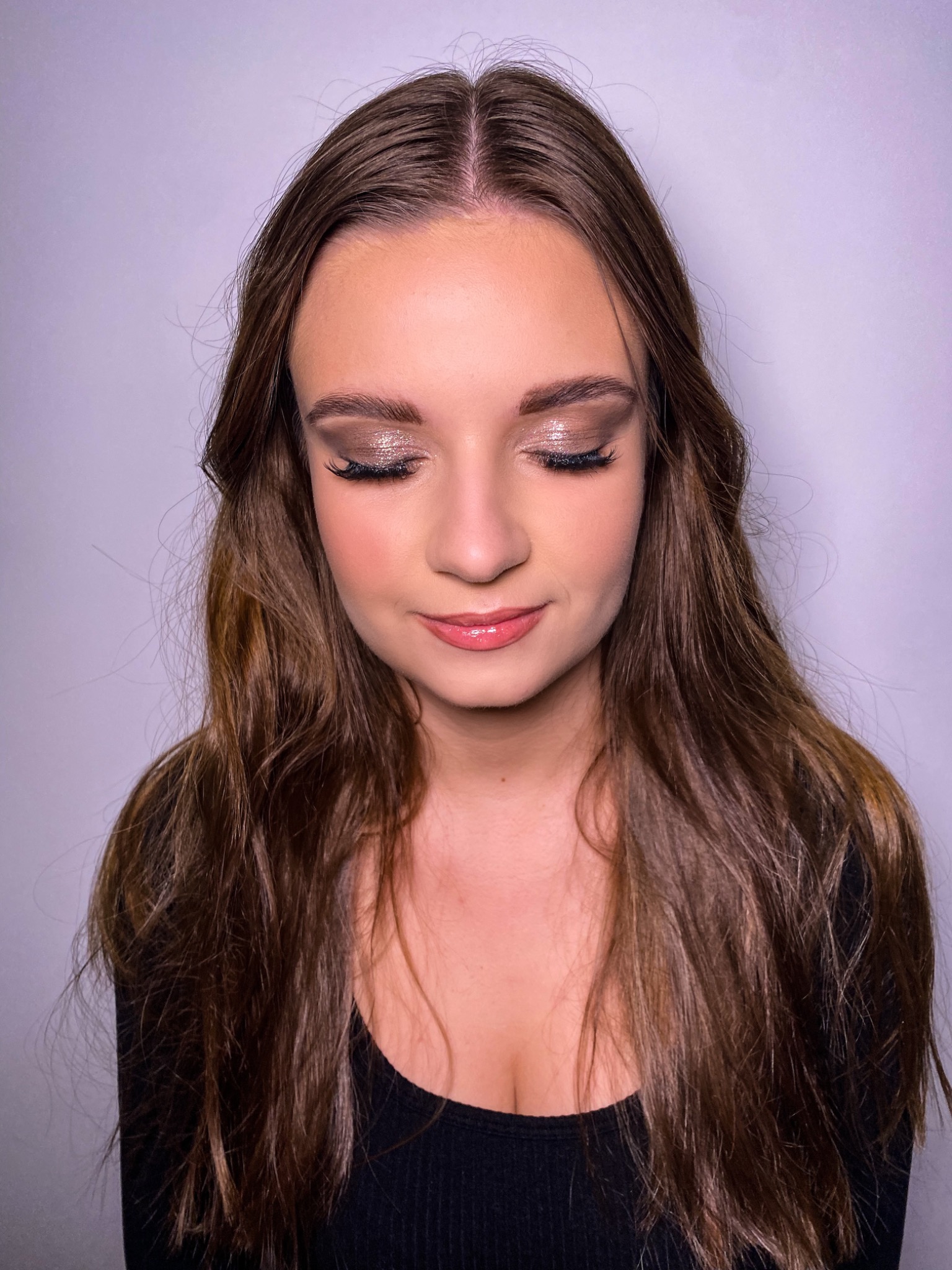 Portret kobiety z zamkniętymi oczami prezentujący makijaż oczu z brokatowymi cieniami i delikatny błyszczyk na ustach, na gładkim, jasnym tle.