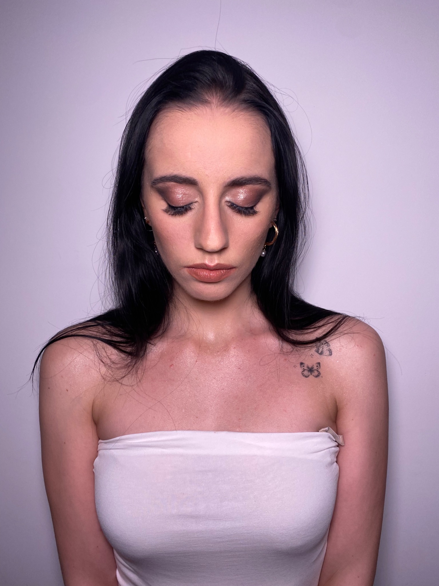 Portret kobiety z ciemnymi włosami i subtelnym, błyszczącym cieniem na powiekach, z widocznymi złotymi kolczykami i tatuażem motyla na ramieniu, pozującej na jasnym tle.