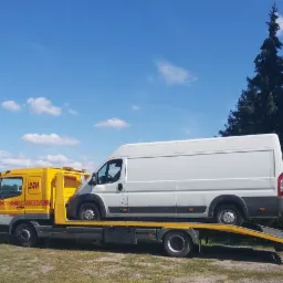 Żółta laweta z napisem 'Pomoc Drogowa 24H' przewożąca białego busa na tle błękitnego nieba i zielonego drzewa.