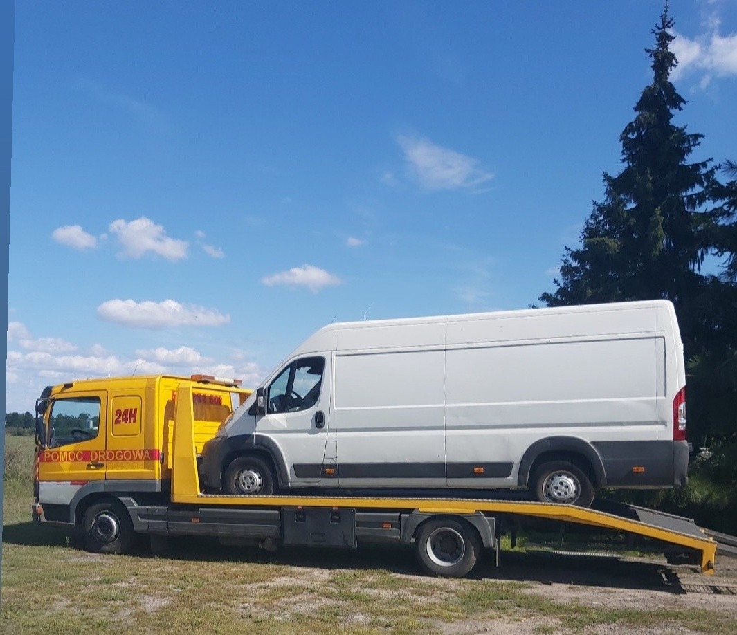 Żółta laweta z napisem 'Pomoc Drogowa 24H' przewożąca białego busa na tle błękitnego nieba i zielonego drzewa.