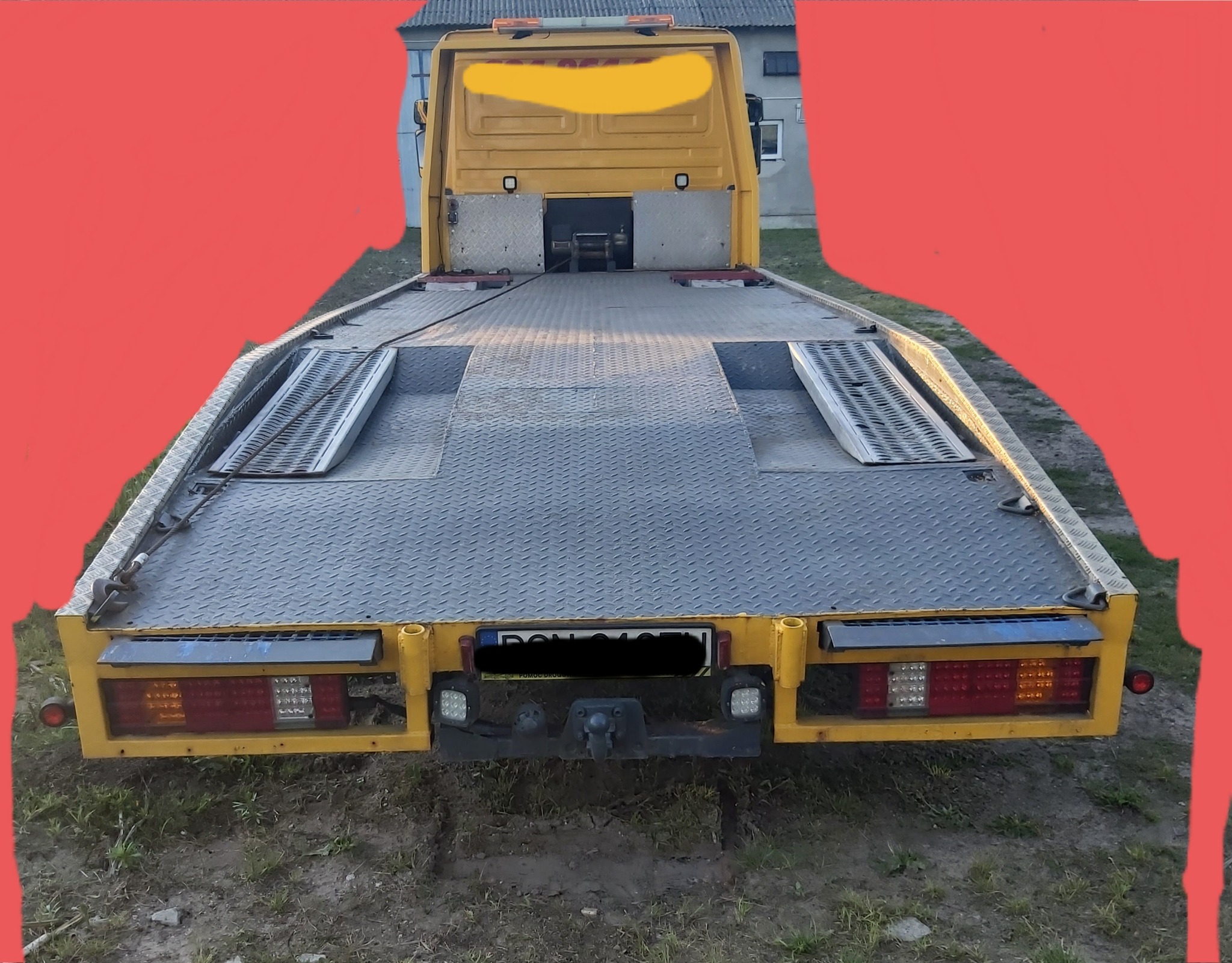 Żółta laweta z metalową platformą, widok z tyłu, z widocznymi światłami, hak holowniczy i fragmentem budynku w tle.