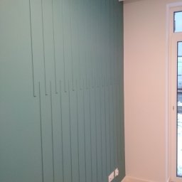 DANIEL GIERASIMIUK - Ściana w kolorze butelkowej zieleni z pionowymi listwami dekoracyjnymi o różnej długości, jasna podłoga, białe ściany i okno w tle.