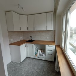 DANIEL GIERASIMIUK - Nowoczesna, jasna kuchnia z białymi szafkami, drewnianym blatem i szarymi marmurkowymi płytkami podłogowymi, widoczne akcesoria kuchenne w otwartej szafce pod zlewem, okno z drewnianym parapetem.