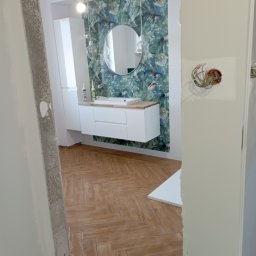 DANIEL GIERASIMIUK - Wykończenie łazienki w stylu nowoczesnym, z drewnopodobną podłogą ułożoną w jodełkę, wiszącą szafką z umywalką na tle tapety w zielone wzory, widoczne niedokończone prace przy ścianie i gniazdku...