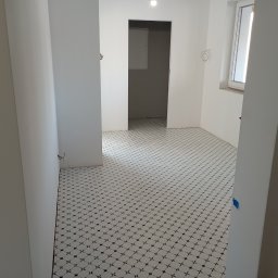 DANIEL GIERASIMIUK - Białe wnętrze w trakcie prac wykończeniowych, widoczne białe ściany, podłoga z białych płytek z ciemnym wzorem, otwarte drzwi i wystające kable ze ściany.