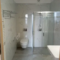 Nowoczesna, jasna łazienka z kabiną prysznicową, wiszącą toaletą i marmurowymi płytkami, wyposażona w uchwyt dla niepełnosprawnych i chromowany wieszak na ręczniki.