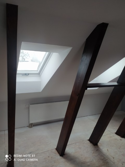 Poddasze z drewnianymi belkami konstrukcyjnymi, okno dachowe z widokiem na zimowy krajobraz, grzejnik pod oknem, przygotowane do dalszych prac wykończeniowych.