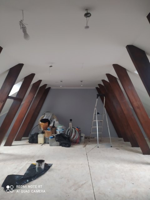Poddasze w trakcie prac przygotowawczych: widoczne drewniane belki konstrukcyjne, zabezpieczona podłoga, drabina, wiadro z farbą i narzędzia malarskie w tle.