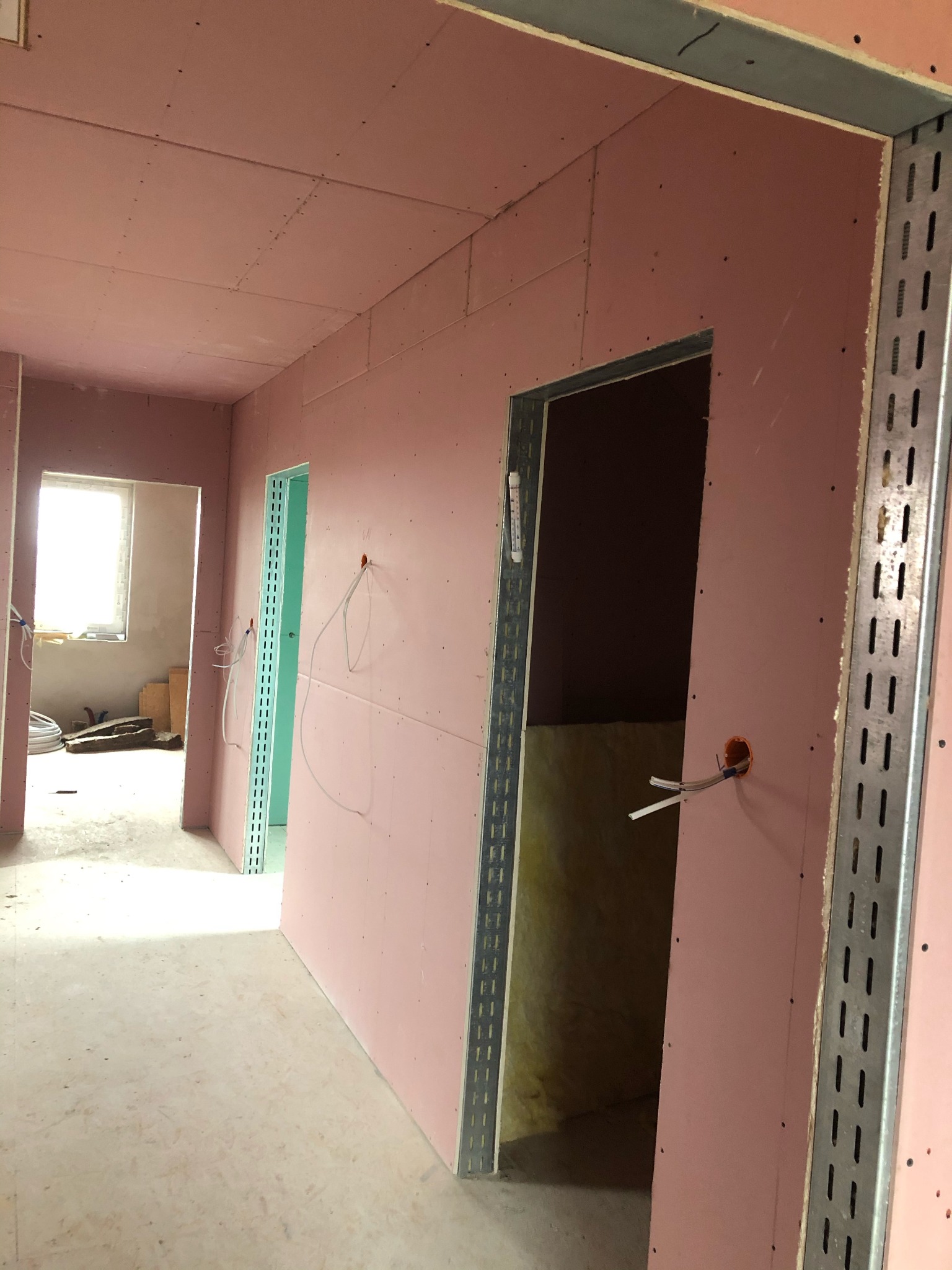 Wnętrze w trakcie prac wykończeniowych: różowe płyty gipsowo-kartonowe na ścianach i suficie, widoczne metalowe profile przy otworach drzwiowych, wystające przewody elektryczne, surowa podłoga...