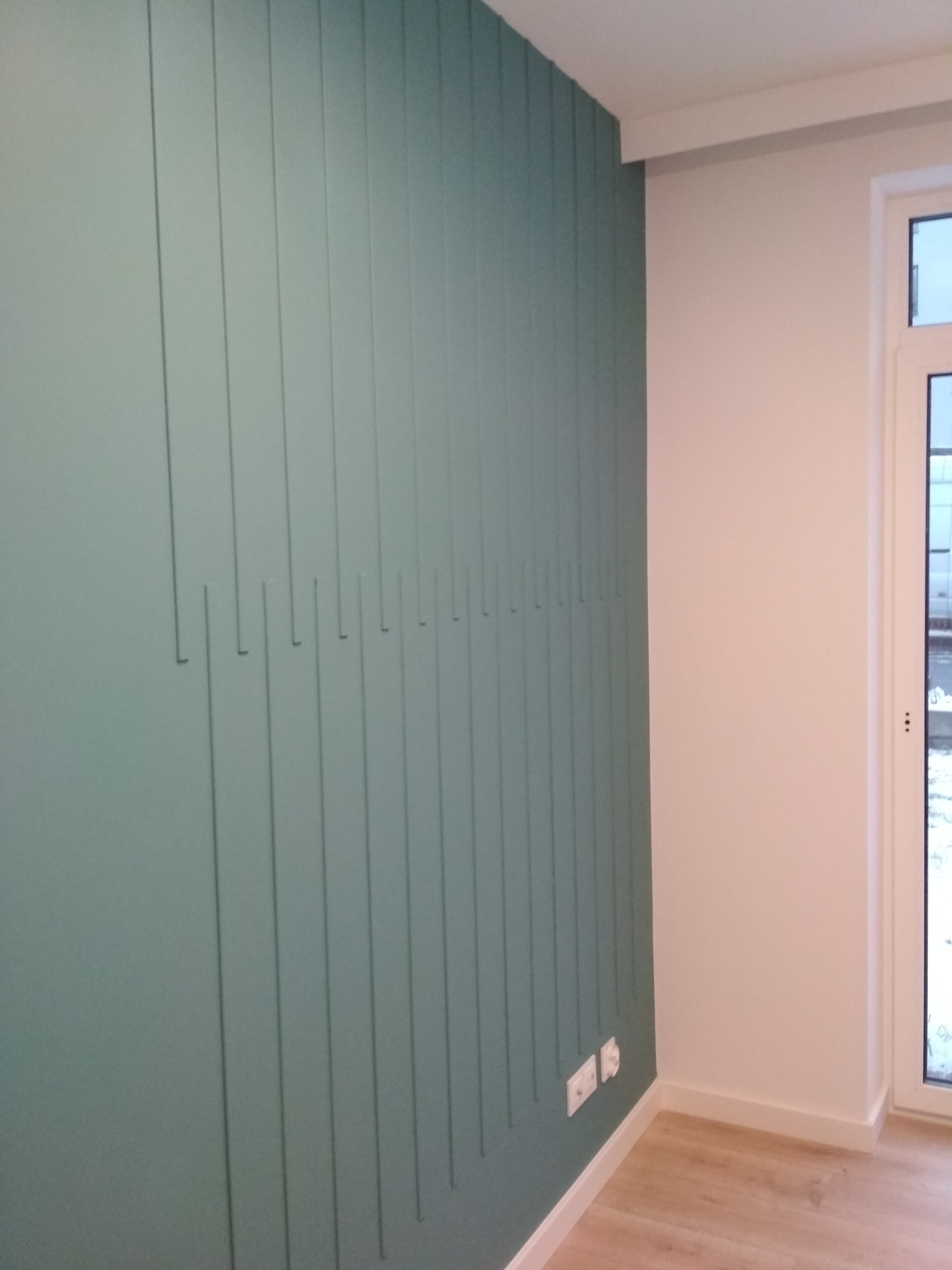 Ściana w kolorze butelkowej zieleni z pionowymi listwami dekoracyjnymi o różnej długości, jasna podłoga, białe ściany i okno w tle.