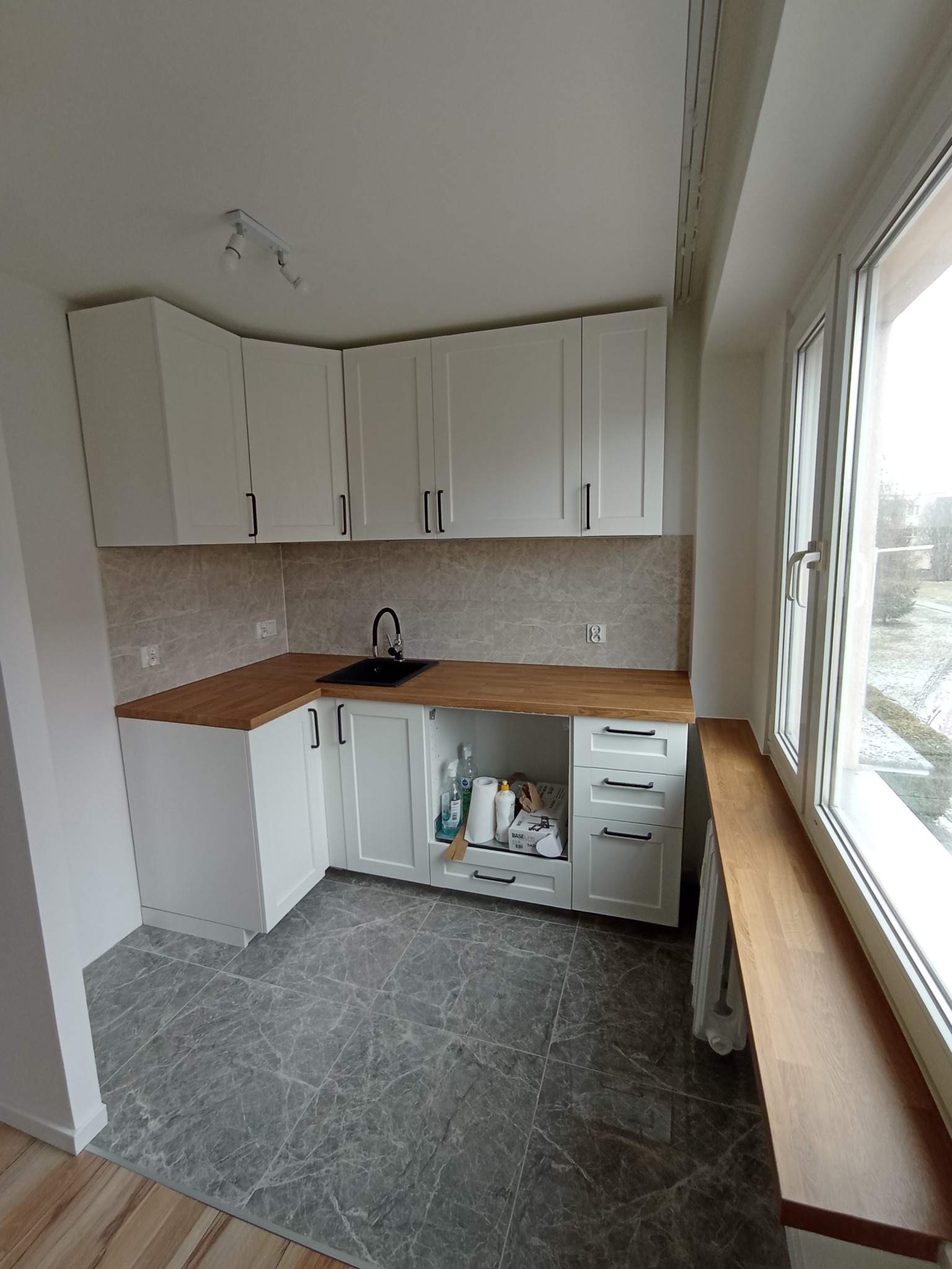 Nowoczesna, jasna kuchnia z białymi szafkami, drewnianym blatem i szarymi marmurkowymi płytkami podłogowymi, widoczne akcesoria kuchenne w otwartej szafce pod zlewem, okno z drewnianym parapetem.