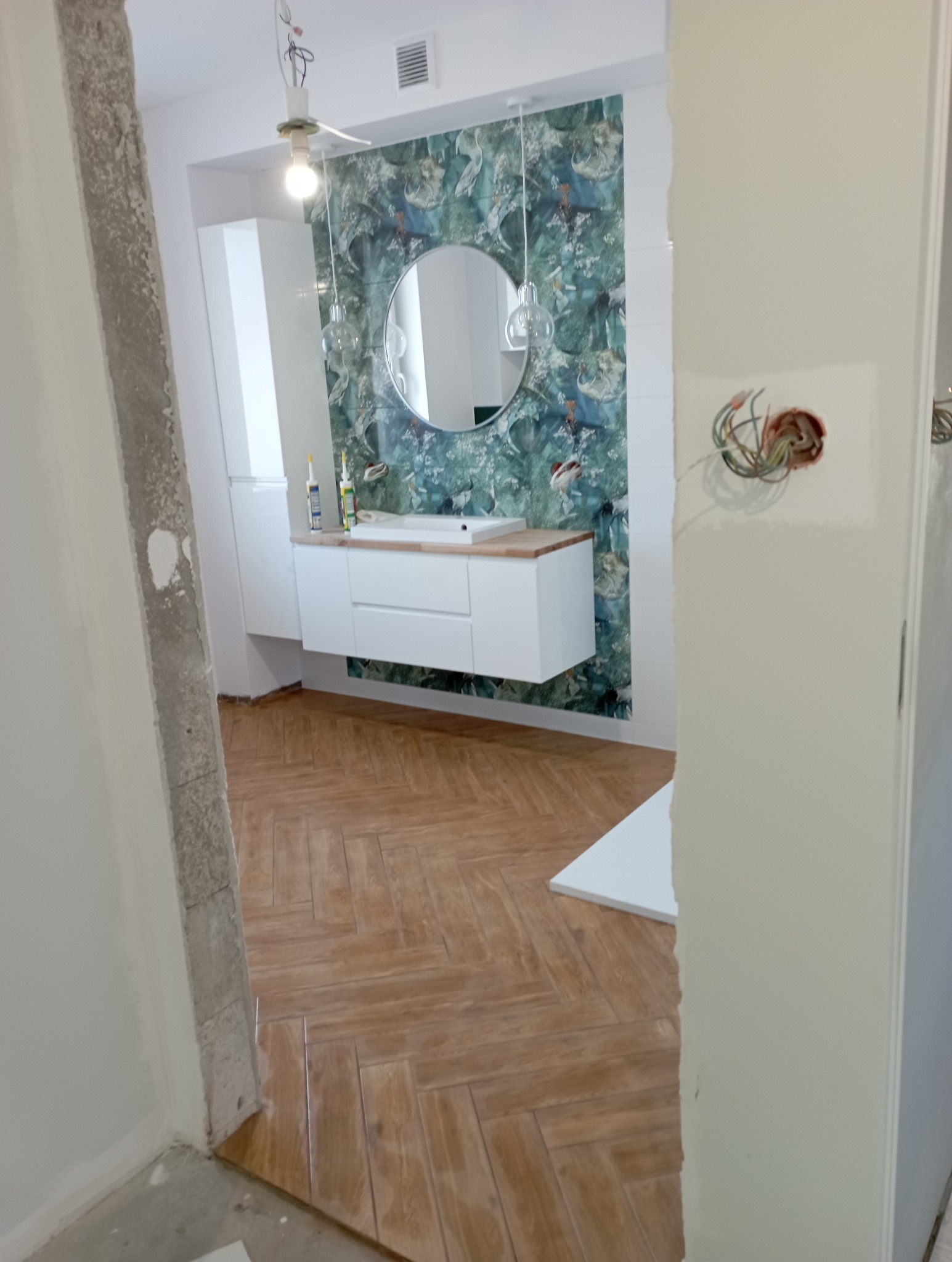 Wykończenie łazienki w stylu nowoczesnym, z drewnopodobną podłogą ułożoną w jodełkę, wiszącą szafką z umywalką na tle tapety w zielone wzory, widoczne niedokończone prace przy ścianie i gniazdku...