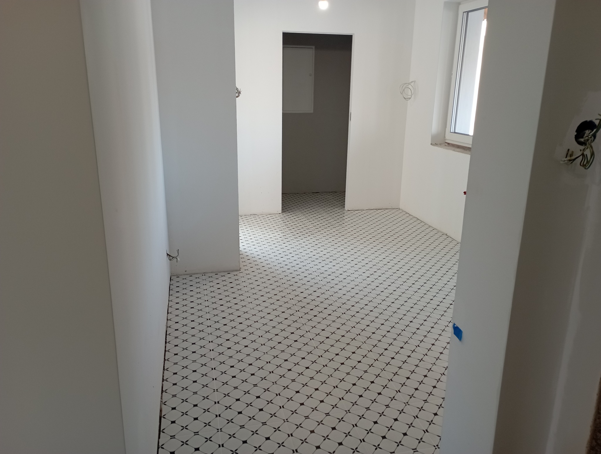 Białe wnętrze w trakcie prac wykończeniowych, widoczne białe ściany, podłoga z białych płytek z ciemnym wzorem, otwarte drzwi i wystające kable ze ściany.