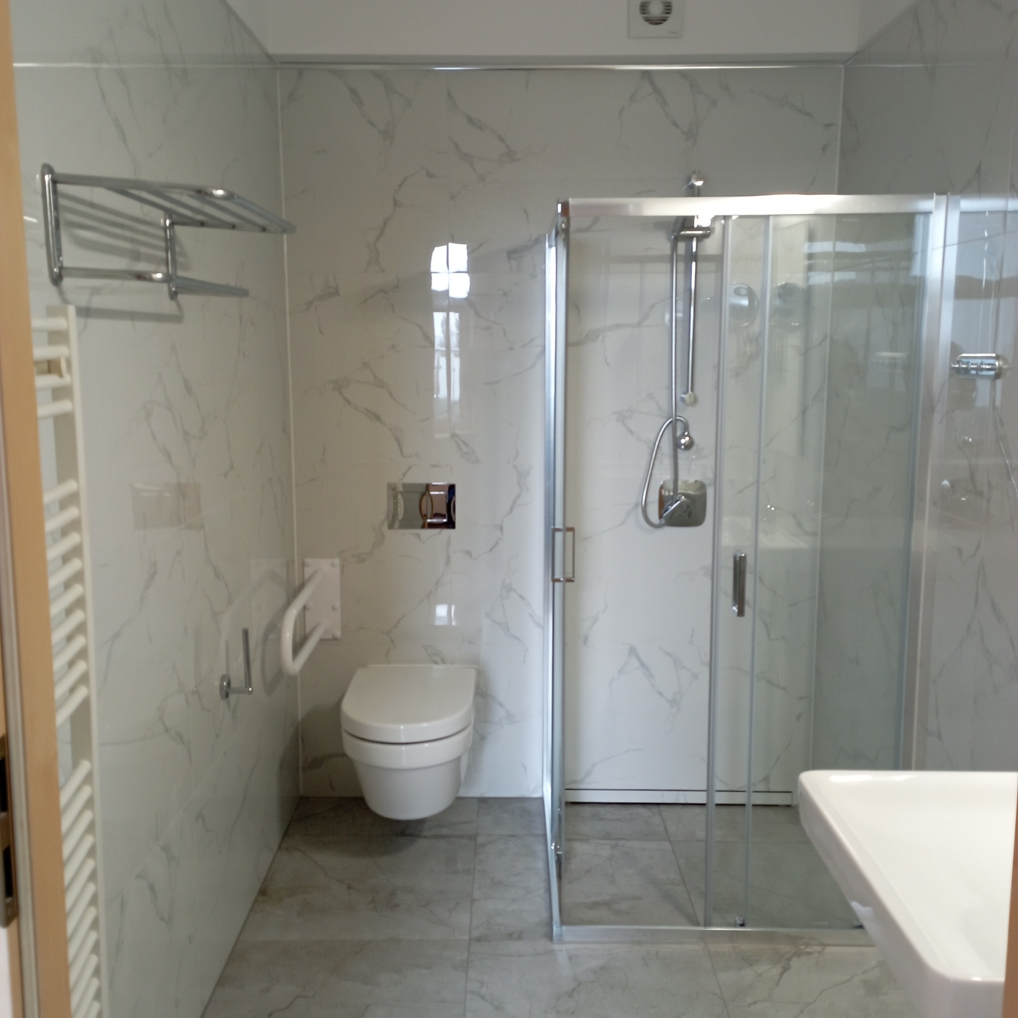 Nowoczesna, jasna łazienka z kabiną prysznicową, wiszącą toaletą i marmurowymi płytkami, wyposażona w uchwyt dla niepełnosprawnych i chromowany wieszak na ręczniki.