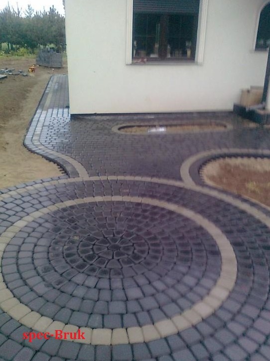 Okrąg z ciemnoszarej kostki brukowej otoczony jaśniejszym pasem, tworzący wzór na podjeździe przed białym budynkiem z oknem i zielonym tłem drzew.