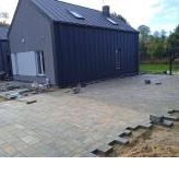 Szara kostka brukowa ułożona częściowo na piasku przed nowoczesnym domem z ciemną elewacją i białymi okiennicami, widoczne niedokończone krawędzie i pojedyncze kostki przygotowane do ułożenia.