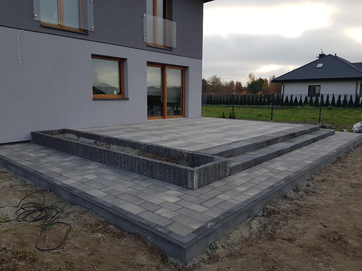 Wykończony taras z kostki brukowej w odcieniach szarości, z niską donicą i schodami, prowadzący do nowoczesnego domu z szarą elewacją i drewnianymi oknami, widok na ogród w tle.