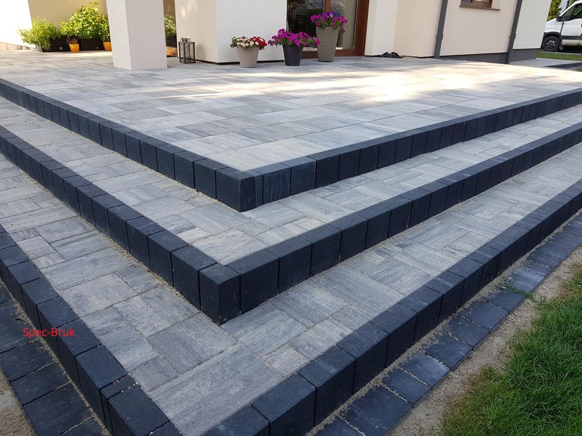 Schody wejściowe do domu w Łodzi wyłożone szarą kostką brukową z ciemnymi obrzeżami, prowadzące do jasnego budynku z kwiatami w donicach.