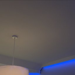 Łukasz Kurgan - Biały sufit z zamontowanym oświetleniem LED w kolorze niebieskim wzdłuż krawędzi oraz lampą wiszącą z abażurem w dolnym lewym rogu.
