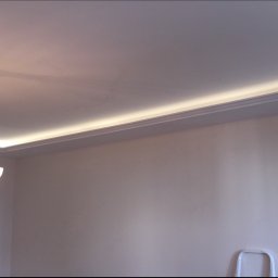 Łukasz Kurgan - Nowoczesny sufit z oświetleniem LED wzdłuż krawędzi, ozdobiony klasycznym żyrandolem z białymi kloszami i rozetą sufitową.