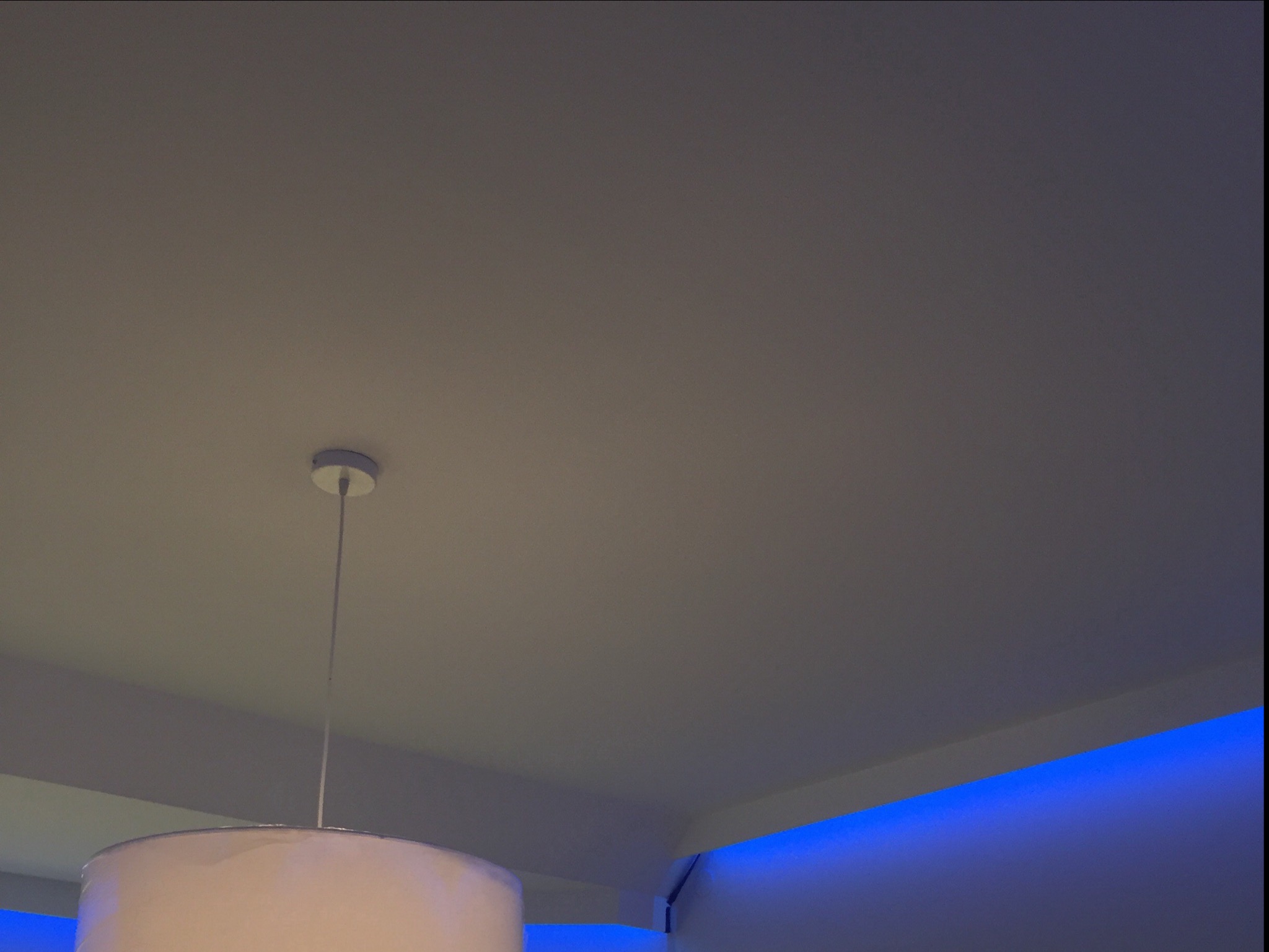Biały sufit z zamontowanym oświetleniem LED w kolorze niebieskim wzdłuż krawędzi oraz lampą wiszącą z abażurem w dolnym lewym rogu.
