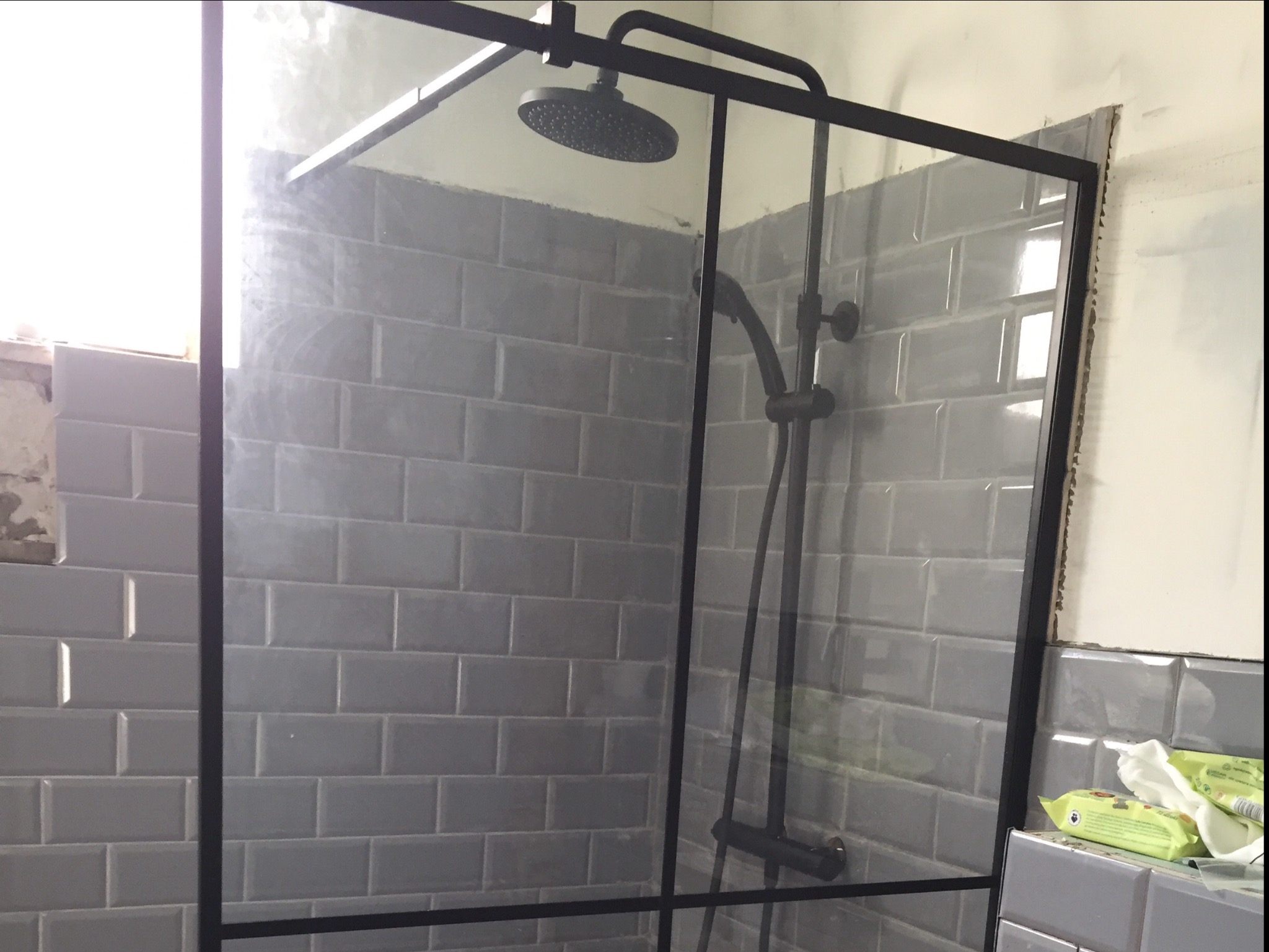 Szklana kabina prysznicowa z czarnymi profilami i deszczownicą, obłożona szarymi, prostokątnymi płytkami imitującymi cegłę, widoczne niedokończone wykończenia ściany i okna.