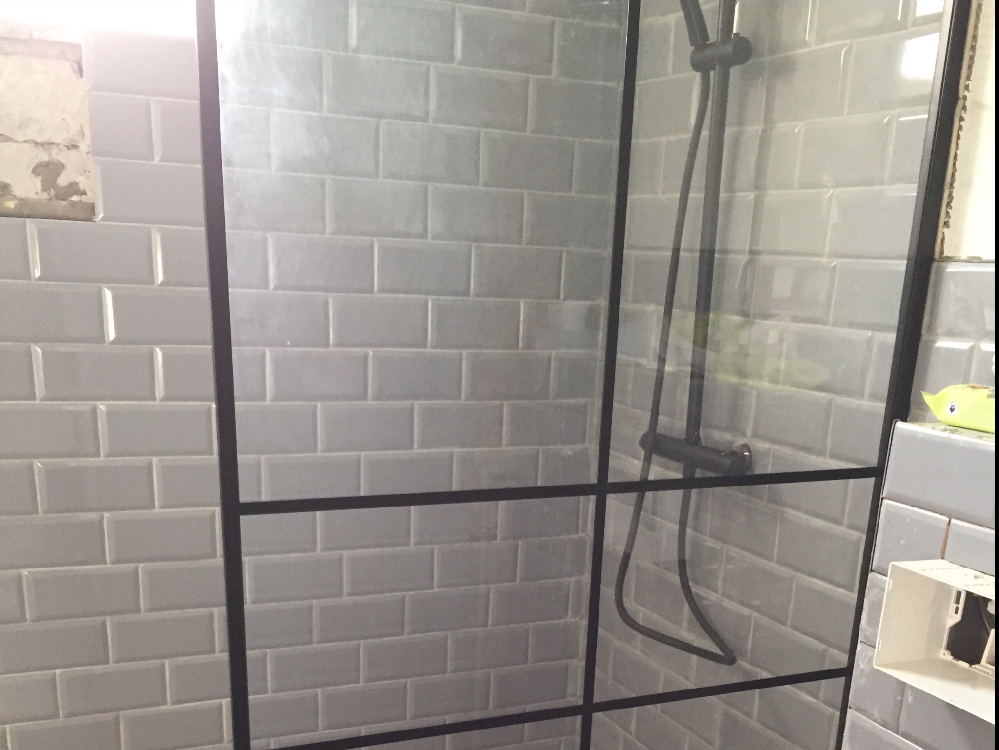 Szklana kabina prysznicowa z czarnymi profilami, szare płytki typu metro na ścianach, widoczna armatura i fragment instalacji elektrycznej w rogu.