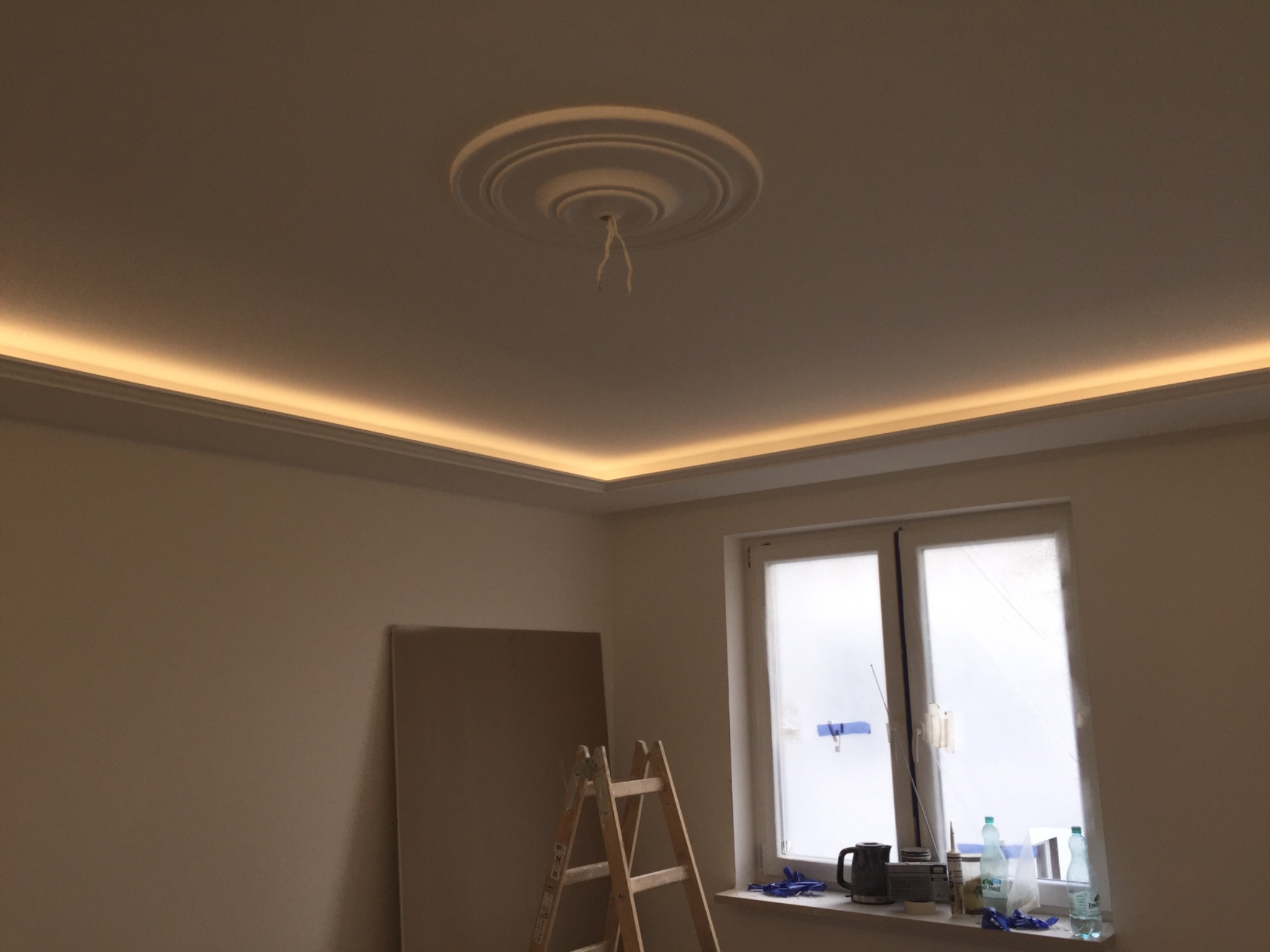 Pokój w trakcie remontu z oświetleniem LED na suficie, widoczna drabina, płyta kartonowo-gipsowa oparta o ścianę oraz okno z przyborami na parapecie.