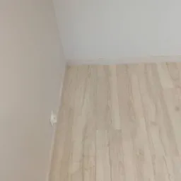 Jasne panele podłogowe ułożone w rogu pokoju, widoczna listwa przypodłogowa i gniazdko elektryczne na ścianie