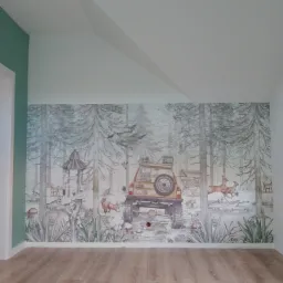 Pokój z fototapetą przedstawiającą leśny krajobraz z samochodem terenowym i zwierzętami, na podłodze panele w odcieniach drewna, ściana po lewej stronie pomalowana na zielono.