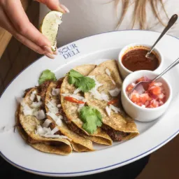 Tacos z wołowiną, cebulą i kolendrą na białym talerzu z niebieskim obramowaniem marki Sour Deli, podawane z salsą i sosem, doprawiane ćwiartką limonki trzymaną w dłoni