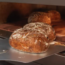 Trzy bochenki chleba rzemieślniczego z dekoracyjnym wzorem nacięć, wyjęte z pieca na metalowej łopacie.