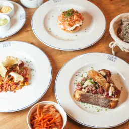 Kompozycja z kilku dań na drewnianym stole, w tym tatar wołowy na grzance, tatar z łososia na bułce, sałatka z marchewki i miseczki z sosami, całość udekorowana gałązkami choinki.