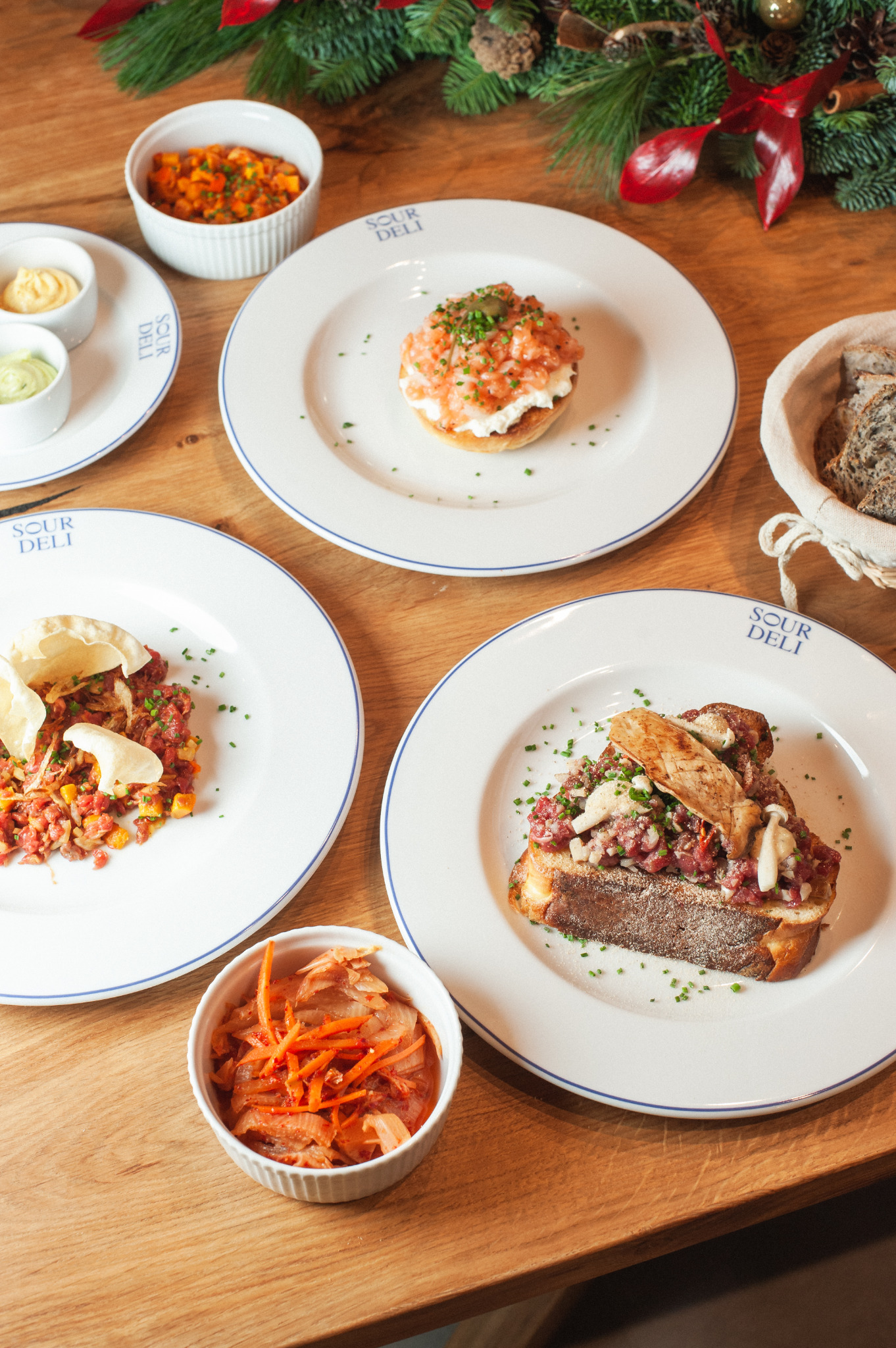 Kompozycja z kilku dań na drewnianym stole, w tym tatar wołowy na grzance, tatar z łososia na bułce, sałatka z marchewki i miseczki z sosami, całość udekorowana gałązkami choinki.