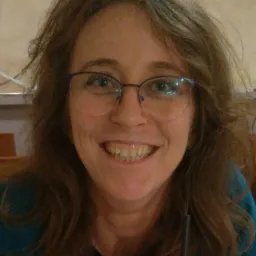 Portret uśmiechniętej kobiety w okularach, z widoczną słomką w szklance, w ciepłym oświetleniu wnętrza.