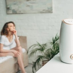 Urocznica.pl - Biały nawilżacz powietrza z beżowymi akcentami na białym stoliku, w tle rozmyta postać kobiety siedzącej na łóżku i roślina doniczkowa.