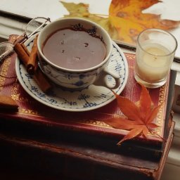 Urocznica.pl - Jesienna kompozycja: filiżanka z napojem, laska cynamonu, ciasteczko, okulary i świeca na stosie starych książek z jesiennymi liśćmi w tle okna.