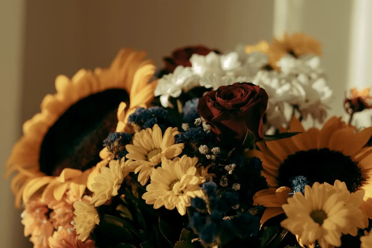 Bukiet mieszanych kwiatów: słoneczniki, czerwona róża, białe i żółte chryzantemy, oraz drobne niebieskie kwiaty, uchwycony w ciepłym, naturalnym świetle.