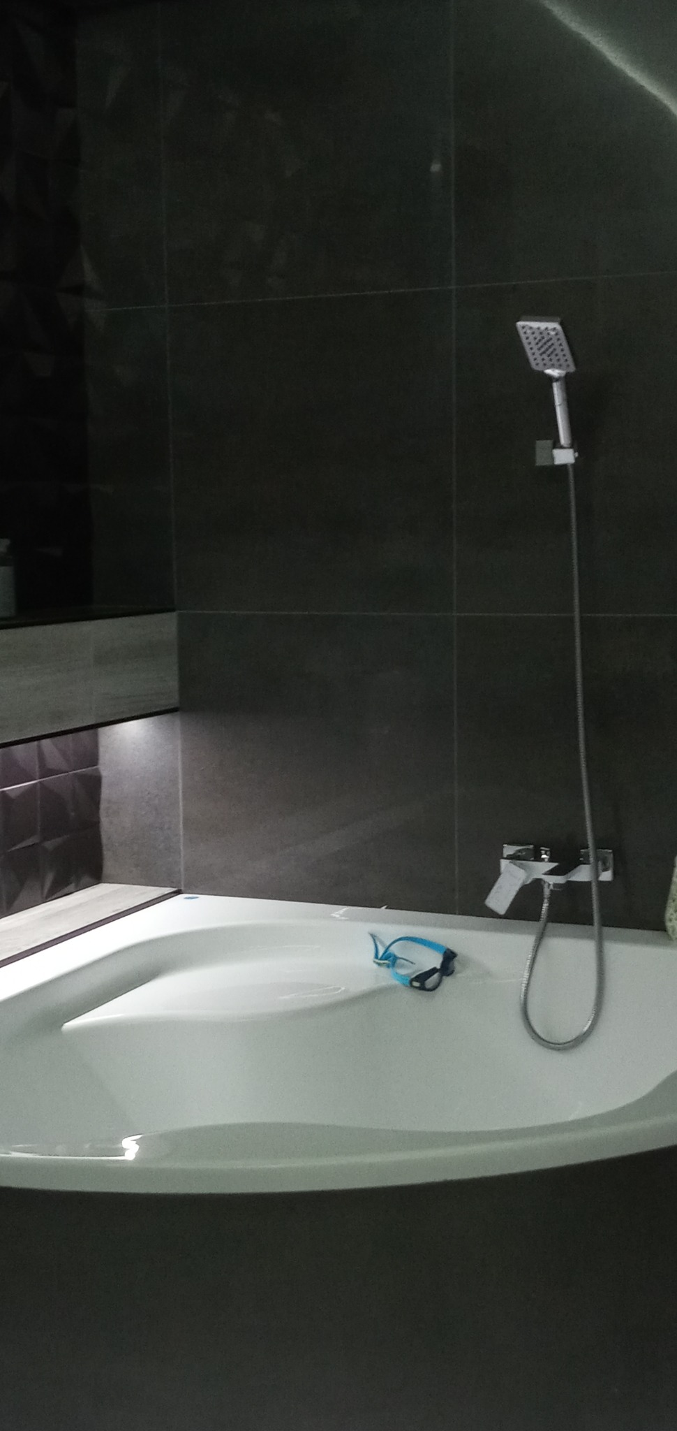 Nowoczesna łazienka z ciemnymi płytkami, białą asymetryczną wanną, słuchawką prysznicową i niebieskimi okularami pływackimi na brzegu wanny.