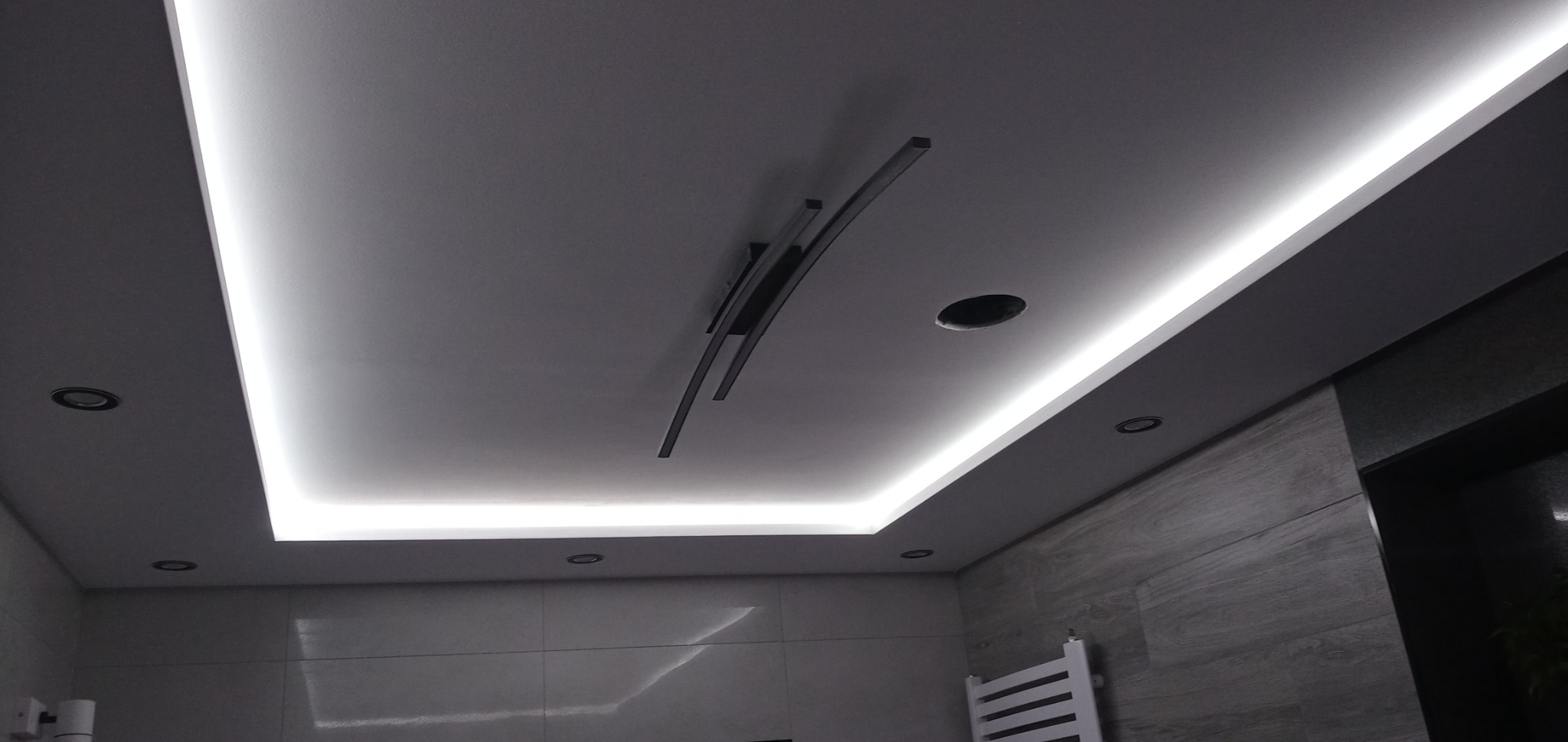 Nowoczesne oświetlenie sufitowe w łazience z listwami LED, dekoracyjnymi czarnymi elementami i punktowymi lampami halogenowymi, widoczne płytki imitujące drewno na ścianach.