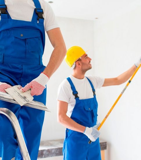 Dwóch mężczyzn w niebieskich kombinezonach i rękawicach roboczych podczas prac wykończeniowych; jeden na drabinie trzyma pacę, drugi maluje ścianę wałkiem na wysięgniku, obaj w jasnym pomieszczeniu.