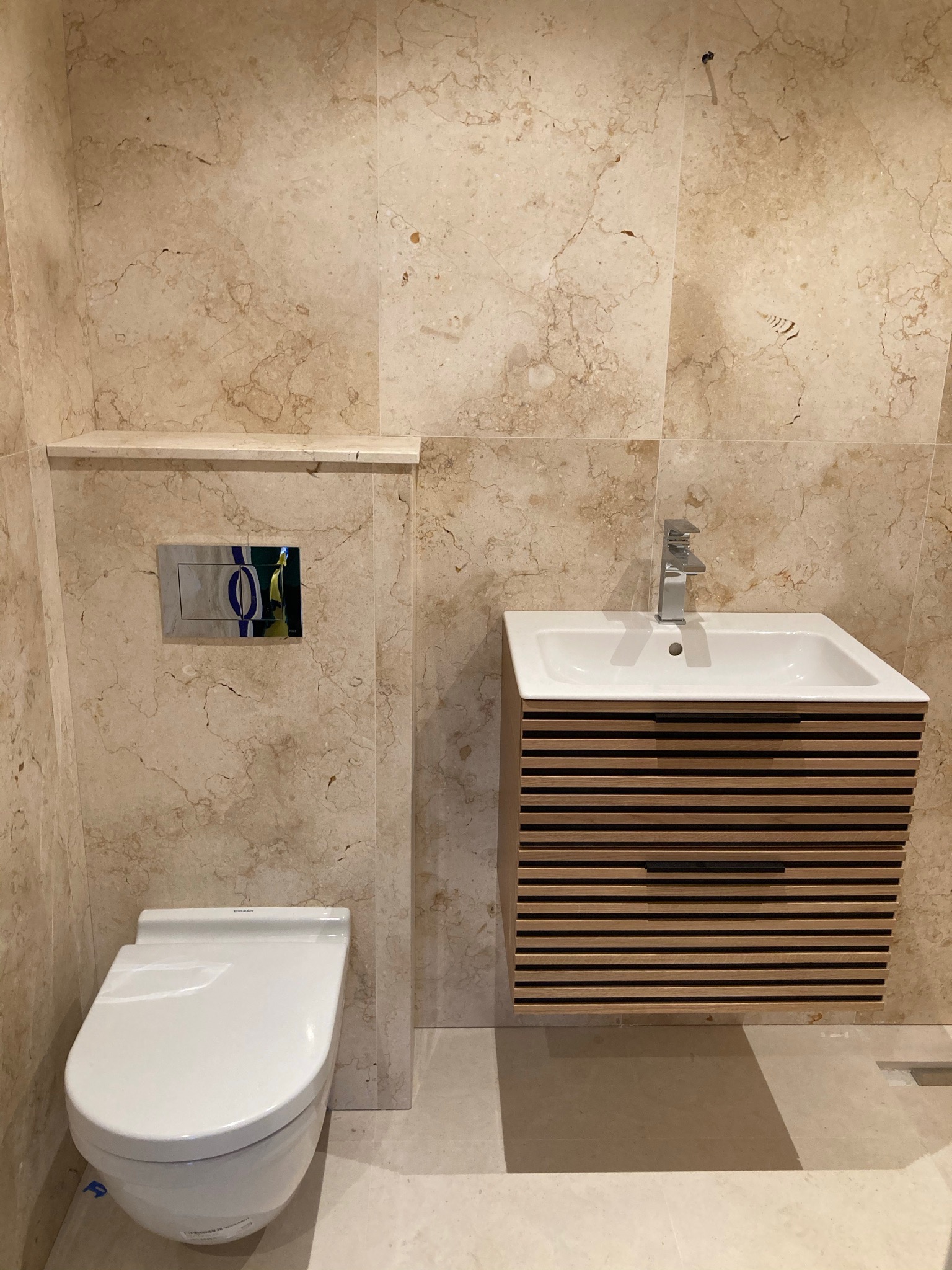 Nowoczesna łazienka z beżowymi płytkami imitującymi marmur, wisząca toaleta, umywalka z szafką z poziomych listewek, chromowana bateria i przycisk spłuczki.